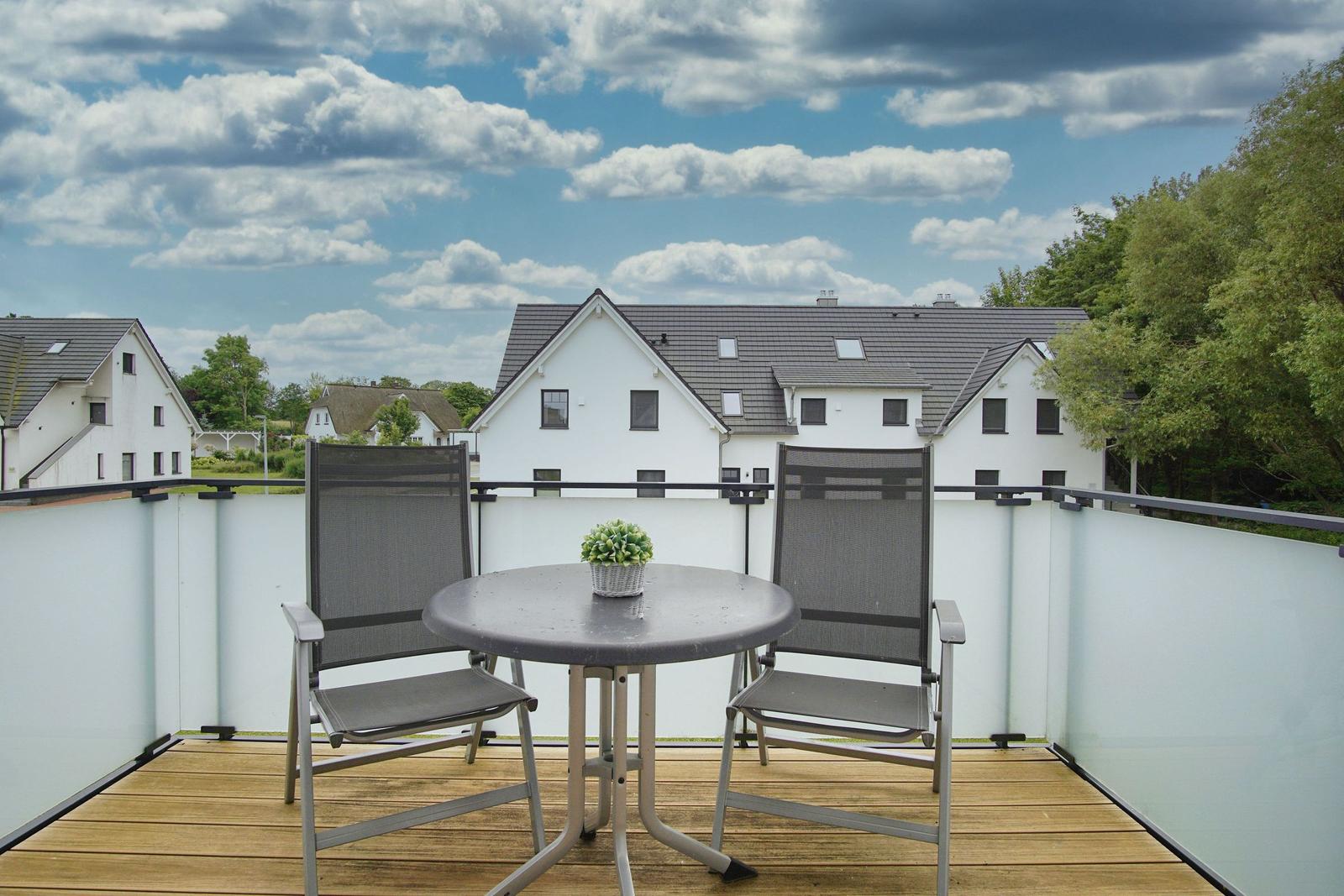 Terrasse mit Tisch und zwei Stühlen, Blick auf weiße Häuser und Bäume unter blauem Himmel.