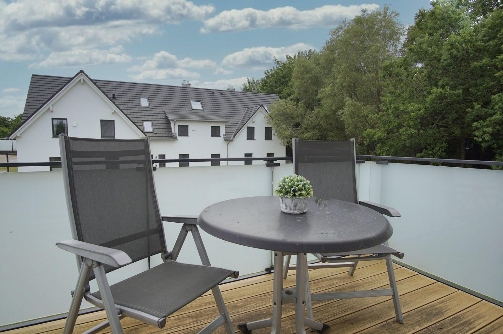 Terrasse mit Tisch und zwei Stühlen. Hintergrund: weißes Haus und Bäume.