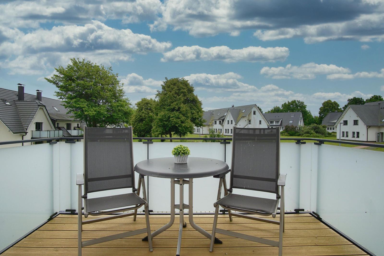 Terrasse mit Tisch und zwei Stühlen. Blick auf Wohnhäuser und Bäume unter bewölktem Himmel.