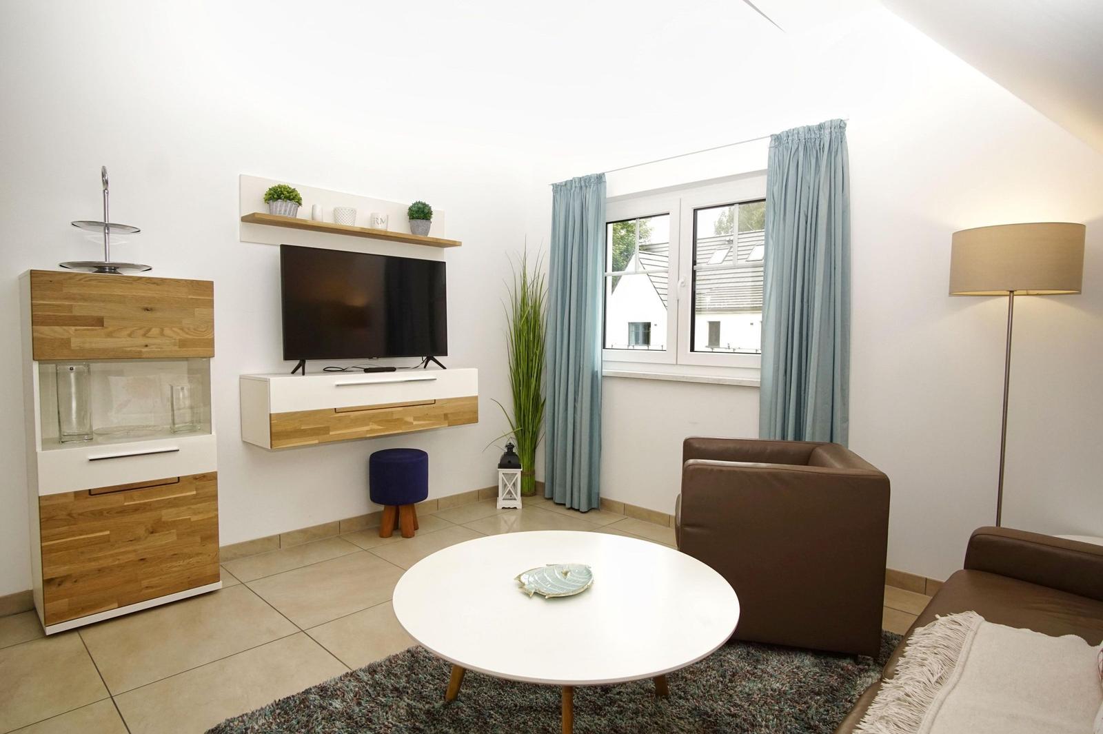 Wohnzimmer mit TV, Sofa, Tisch und Fenster mit Vorhängen.