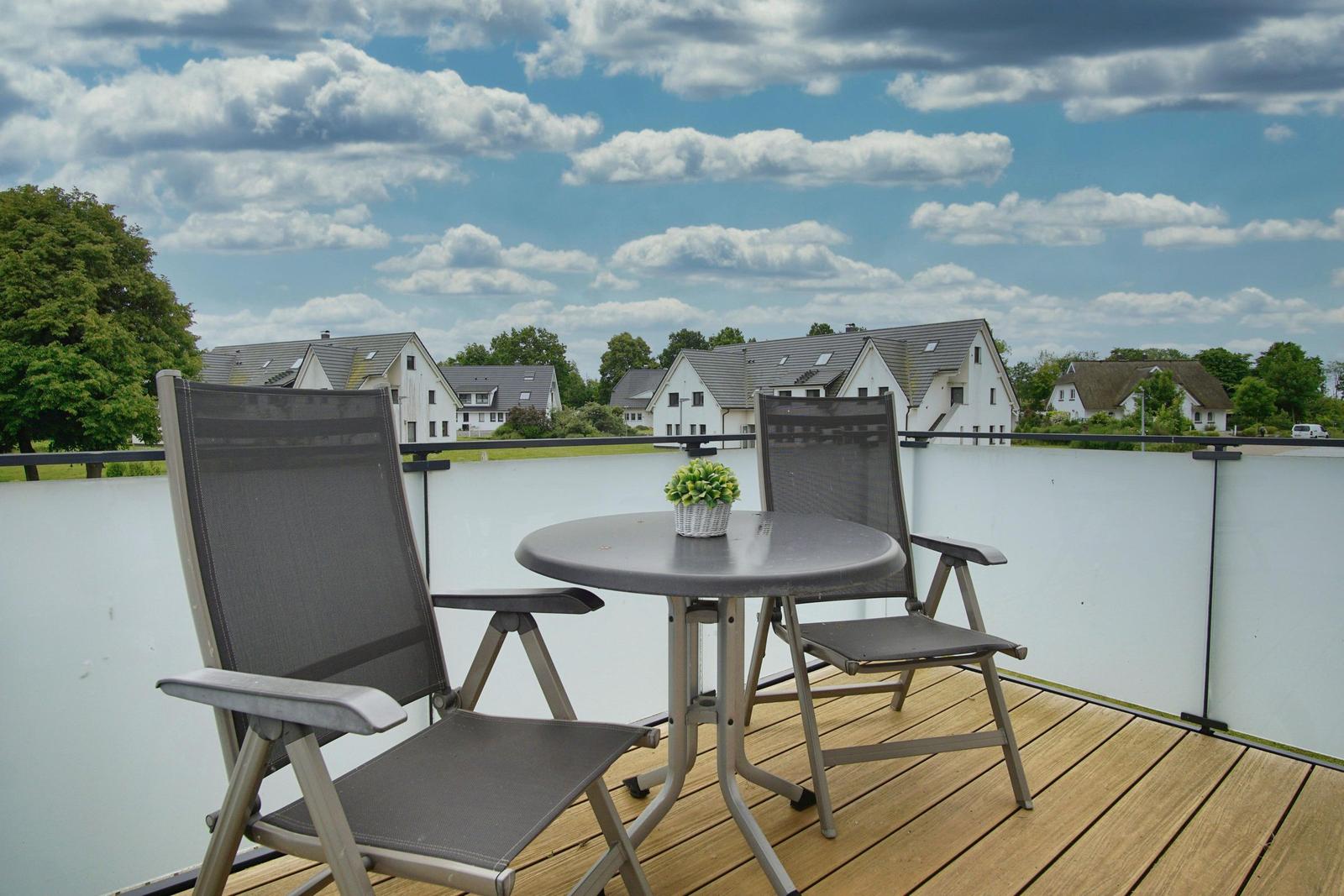 Terrasse mit Tisch und zwei Stühlen. Blick auf Wohnhäuser und Himmel mit Wolken.