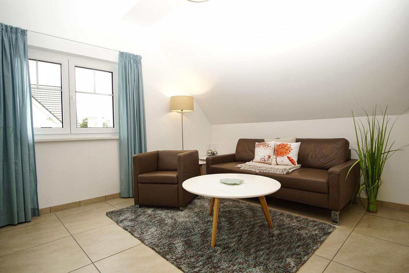 Wohnzimmer mit braunem Sofa, Sessel, runden Tisch und grüner Pflanze.