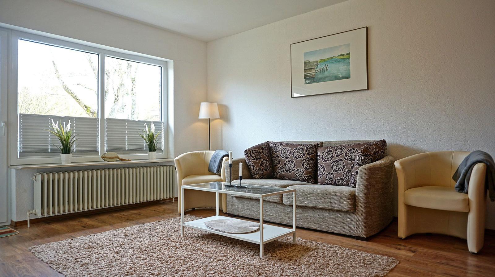 Wohnzimmer mit Sofa, zwei Sesseln, Couchtisch und Fenster mit Jalousien.