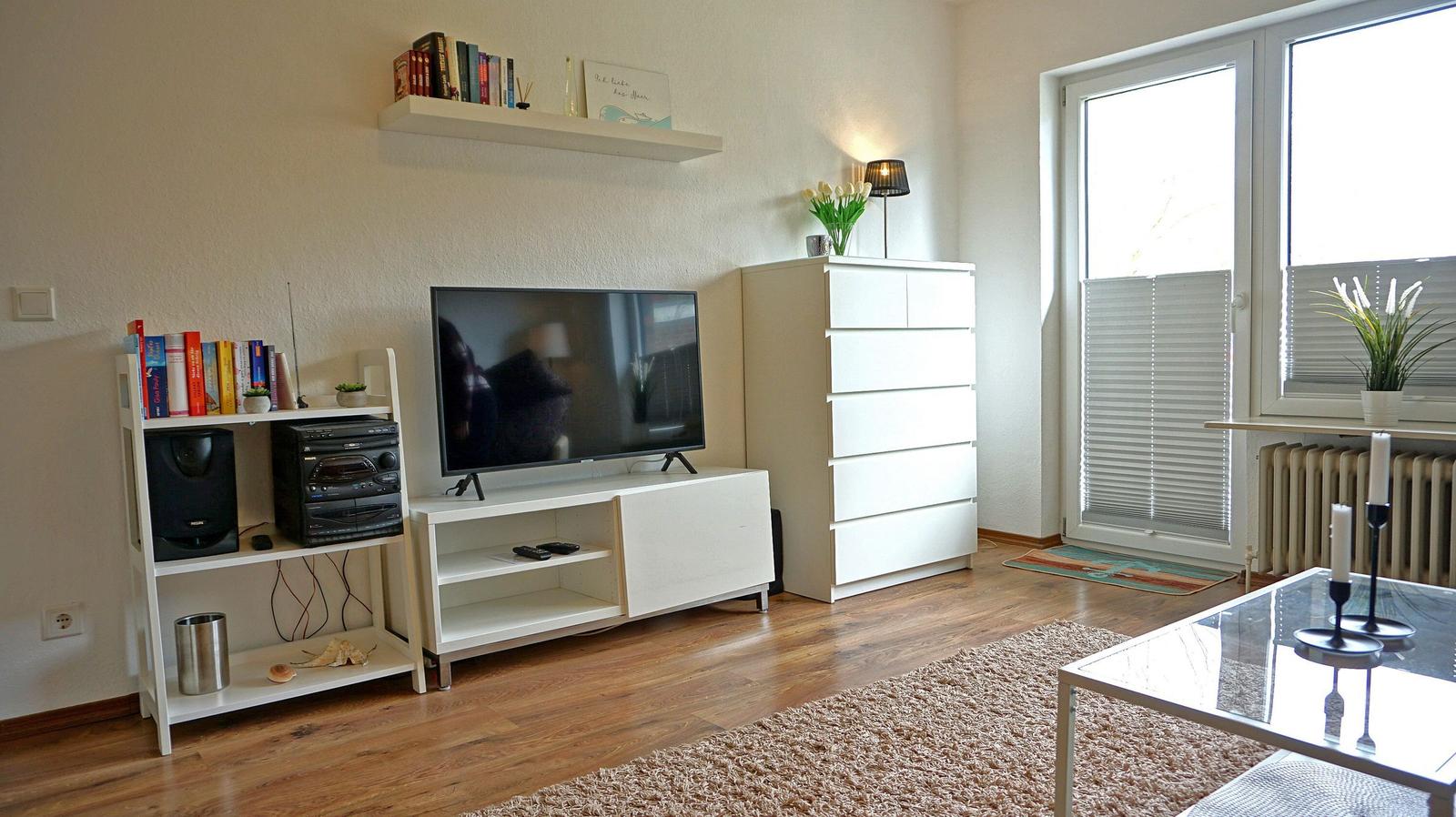 Wohnzimmer mit Fernseher, Regalen, Schrank und Glas-Tisch. Holzboden und Fenster mit Jalousie.