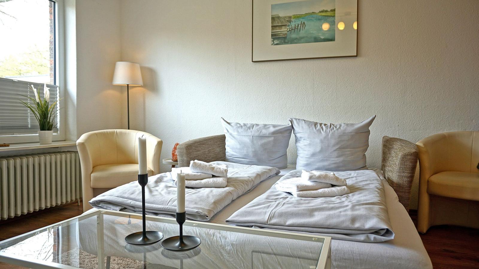 Schlafzimmer mit Bett, zwei Sesseln und Glas-Tisch. Fenster, Wandbild und Stehlampe sichtbar.