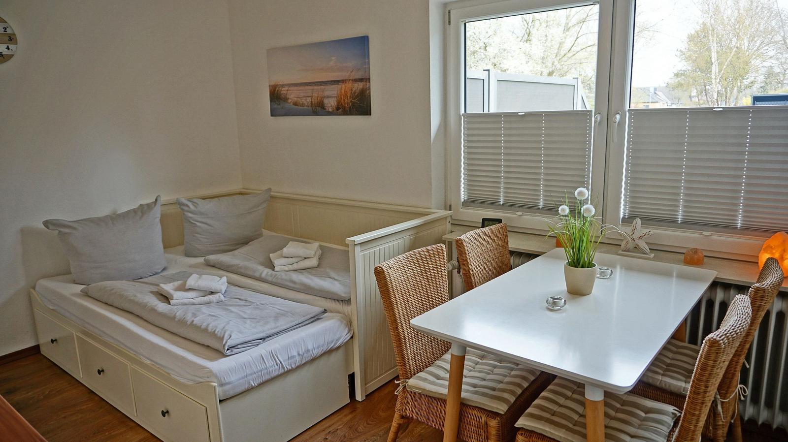 Zimmer mit Schlafgelegenheit und Esstisch mit Stühlen. Fenster mit Jalousien und Wandbild.