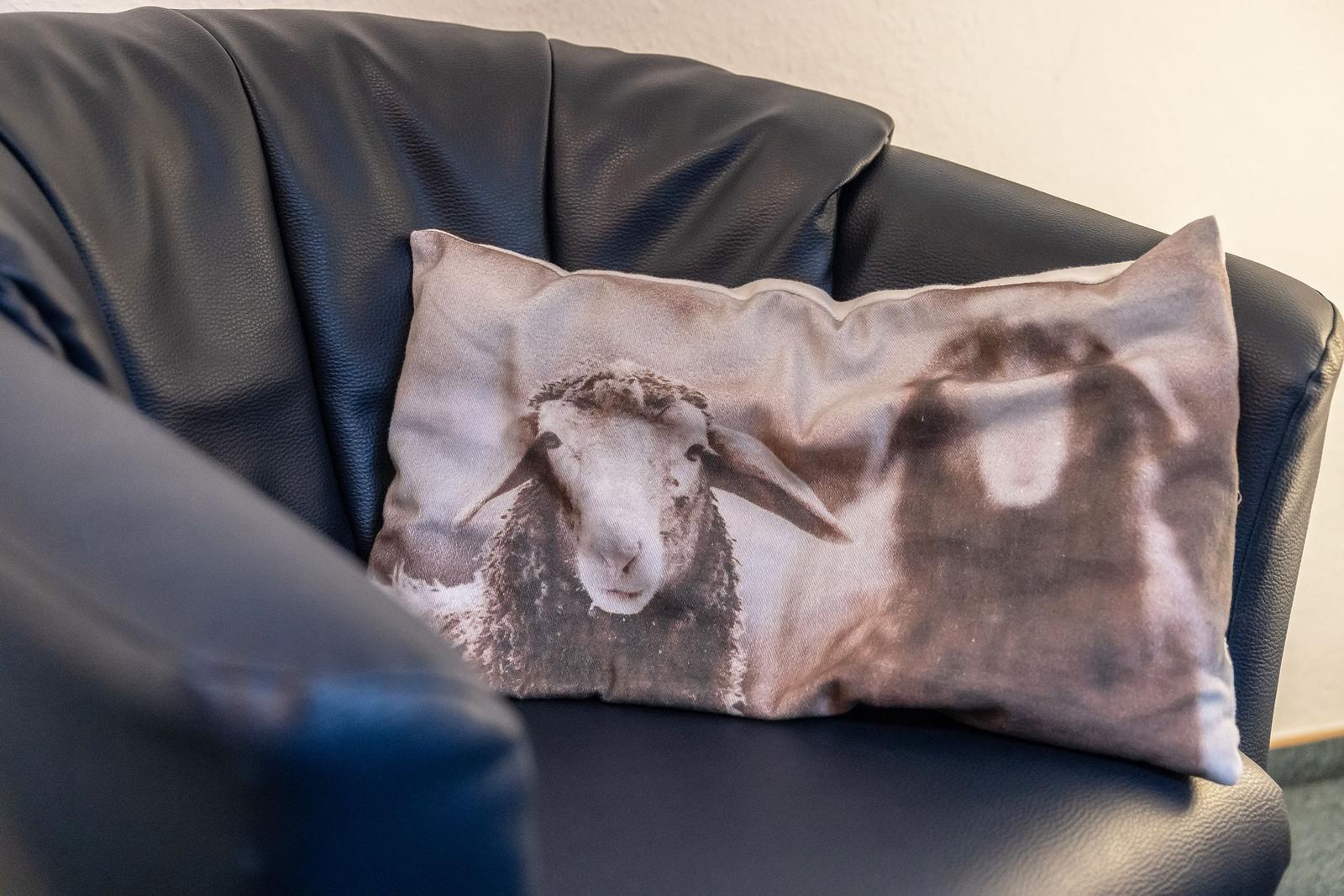Ein Kissen mit Schafmotiv liegt auf einem schwarzen Ledersofa.