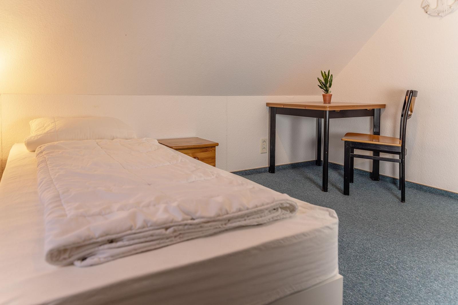 Ein Schlafzimmer mit Bett, Nachttisch, Tisch und Stuhl. An der Wand hängt eine Lampe.