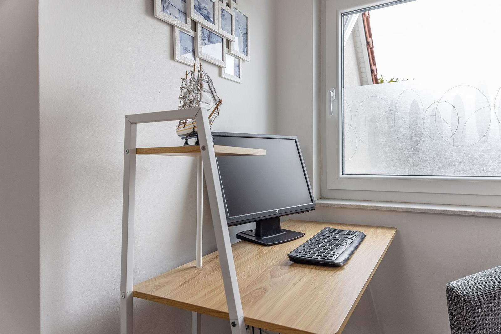 Schreibtisch mit Monitor und Tastatur neben Fenster mit Dekorationsbildern und Segelschiffmodell.
