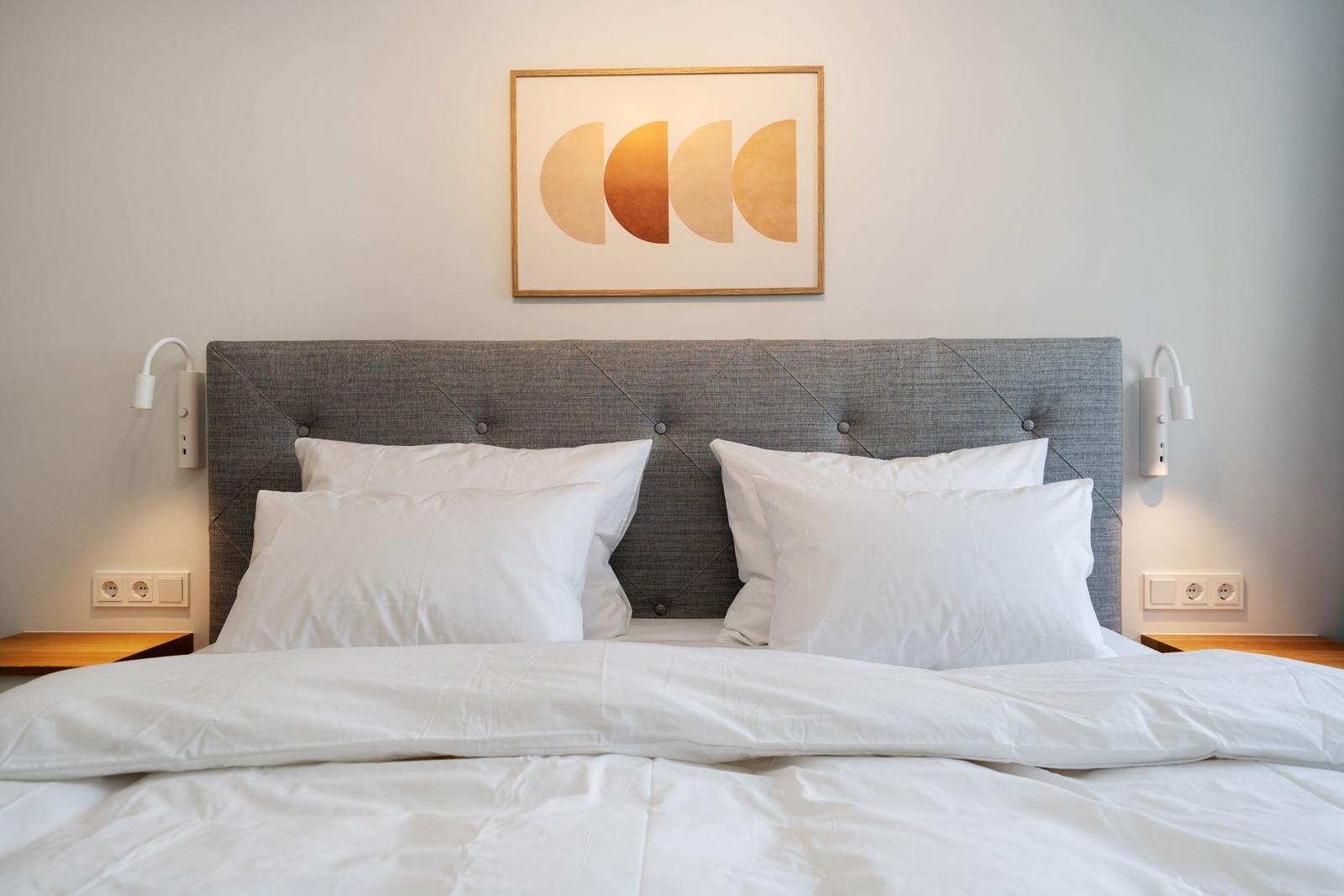 Doppelbett mit weißer Bettwäsche, grauem Kopfteil und Wandbild mit abstraktem Motiv.