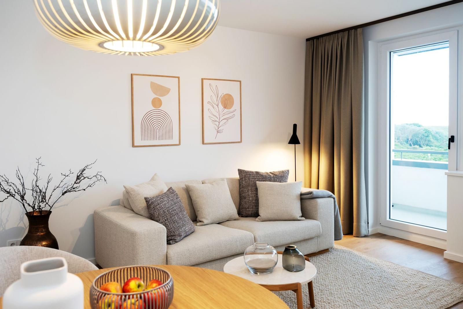 Wohnzimmer mit Sofa, Tisch und Fenster zum Balkon. Wandbilder und Deckenleuchte sichtbar.