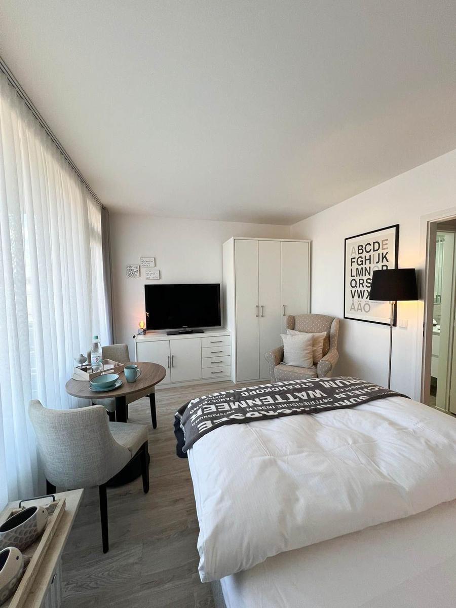 Schlafzimmer mit Bett, TV, Schrank und Sitzbereich. Fenster mit weißen Vorhängen.