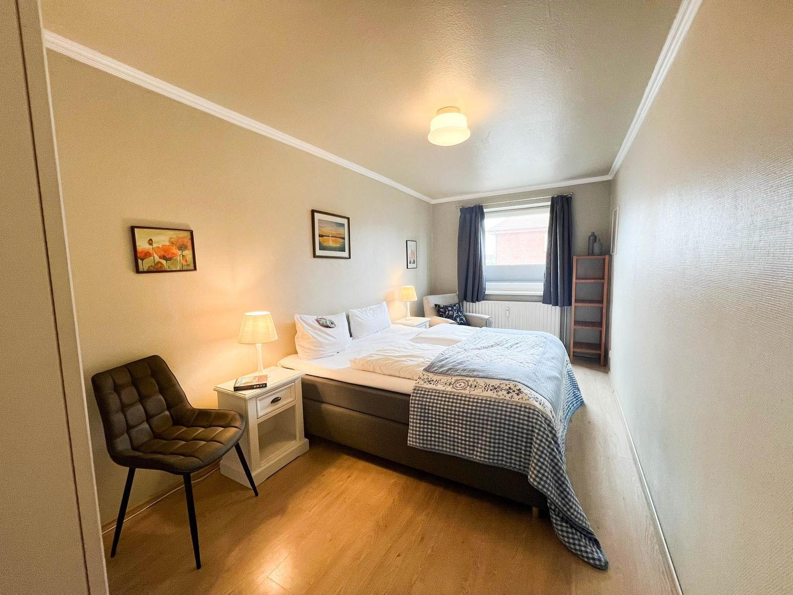 Schlafzimmer mit Bett, Nachttisch, Stuhl und Fenster mit Vorhängen.