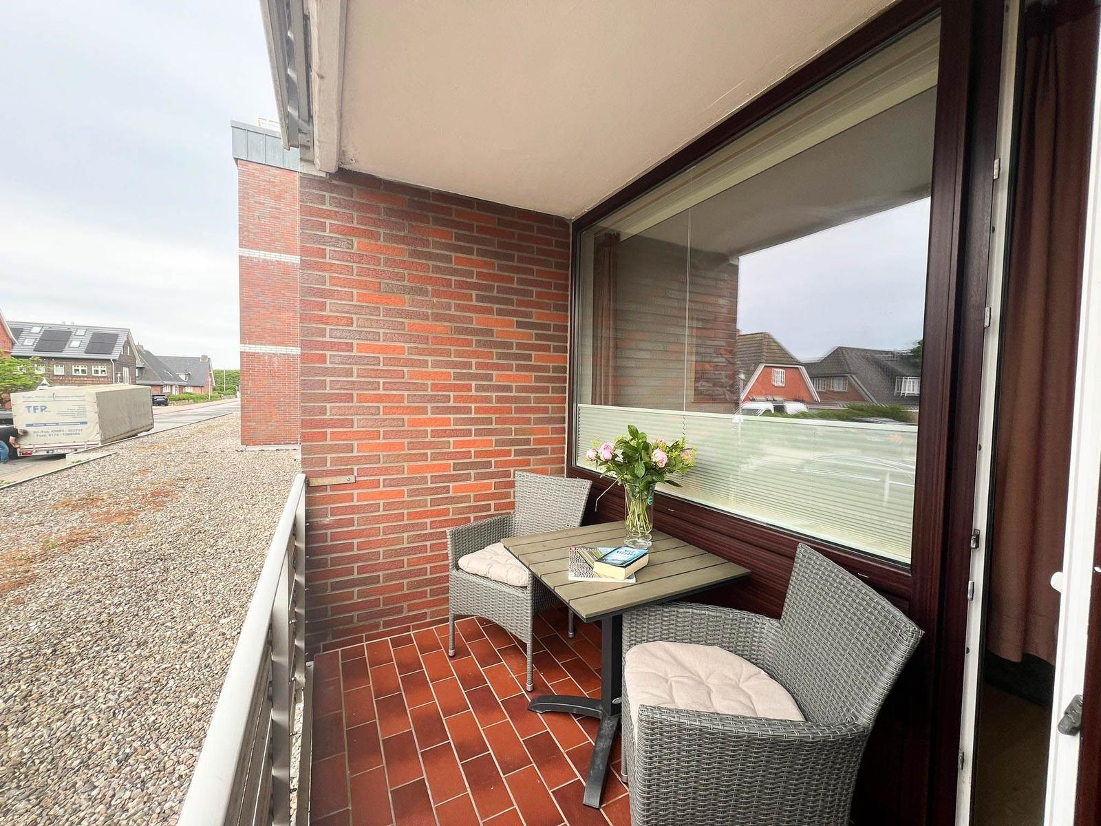 Terrasse mit Tisch und zwei Stühlen, roter Fliesenboden, Blick auf Straße und Häuser.