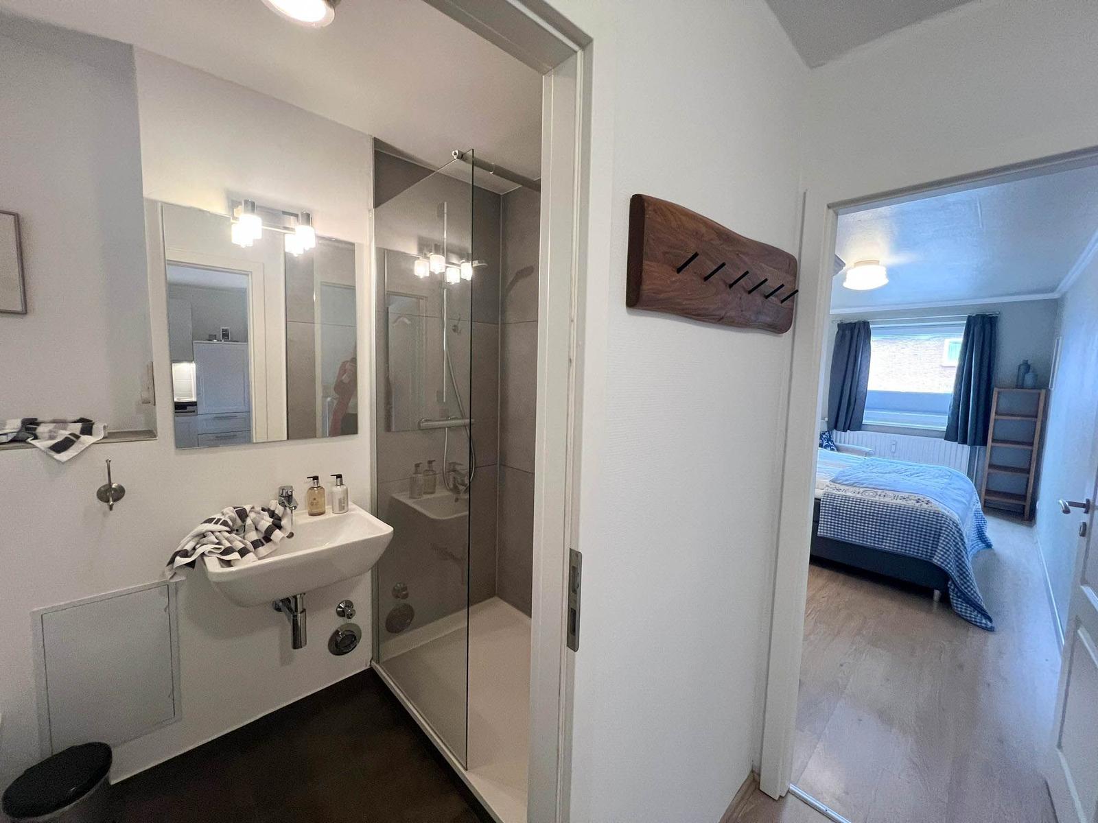 Badezimmer mit Dusche und Waschbecken. Im Hintergrund Schlafzimmer mit Bett.