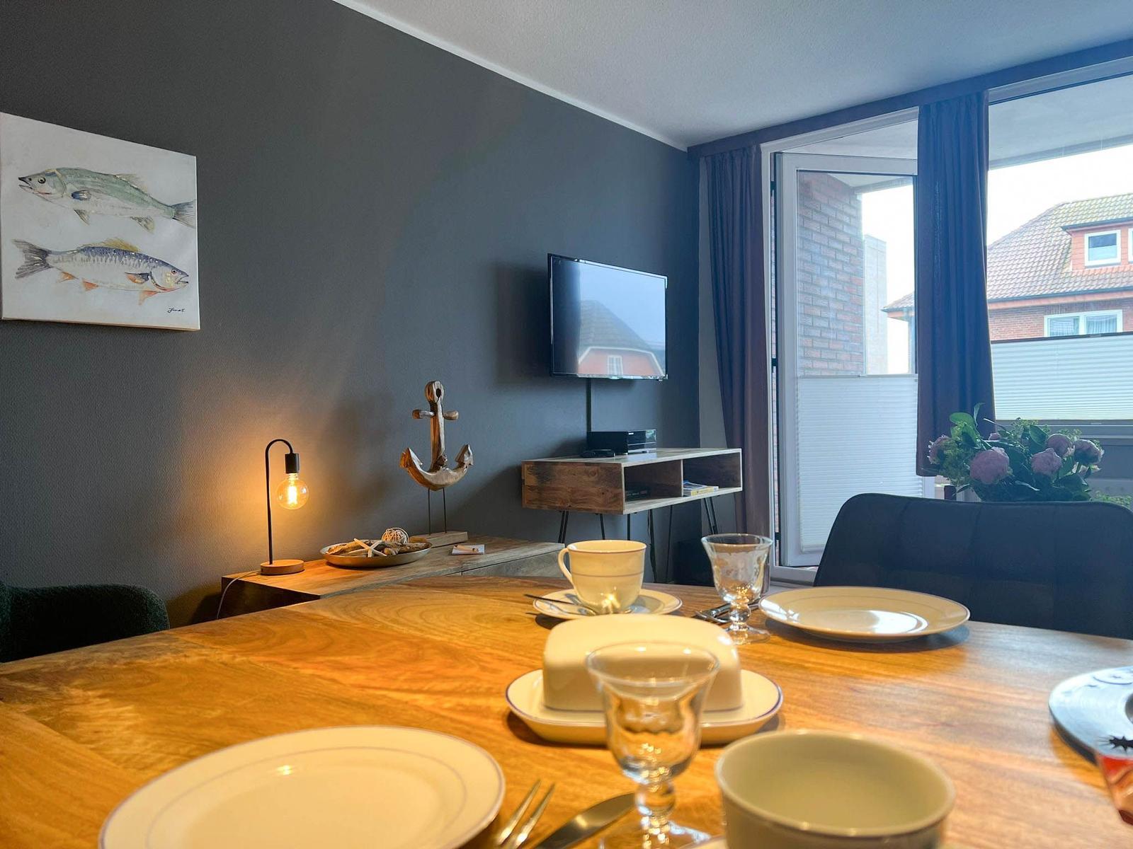 Esstisch mit Geschirr, Wandbild mit Fischen und TV. Blick auf Balkon mit Blumen.