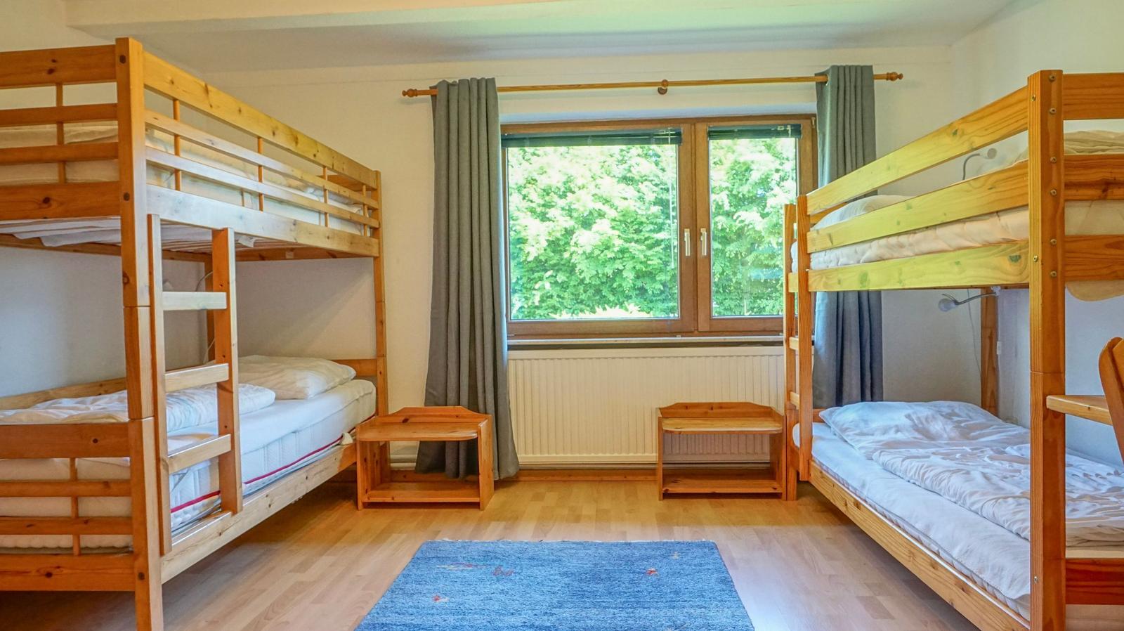Room with two wooden beds, window with view of trees and wooden floor with rug.
