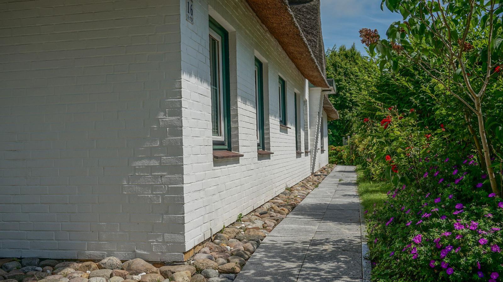 White house wall with green window frames and stone path beside flower beds.