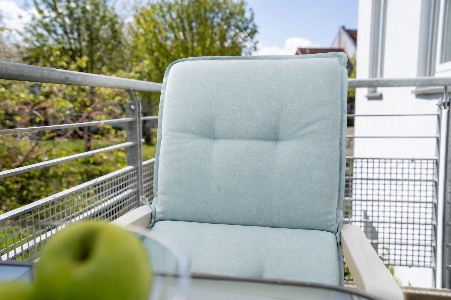 Ein hellblauer Sitzkissen auf einem Balkon mit Metallgeländer und Blick auf Grün.