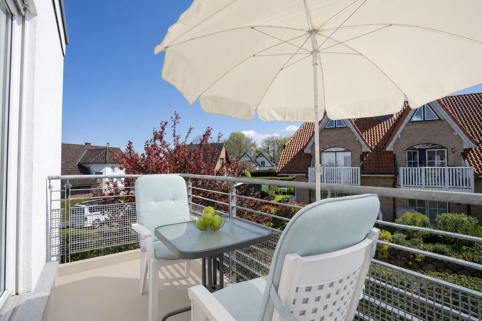 Balcony with table, two chairs, and umbrella. View of houses and trees.
