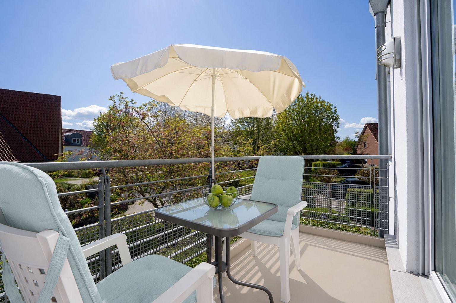 Balkon mit Tisch, zwei Stühlen und Sonnenschirm. Blick auf Bäume und Häuser.