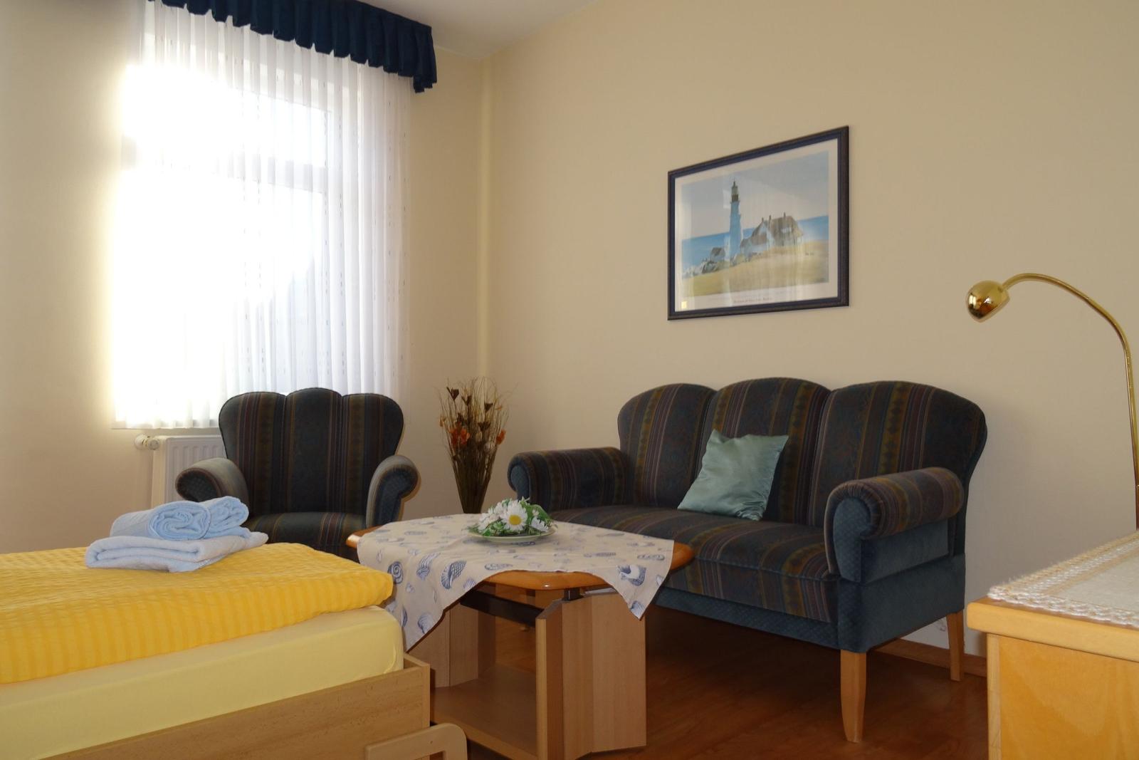 Wohnzimmer mit Sofa, Sessel, Tisch und Fenster. Wandbild und Stehlampe sind sichtbar.