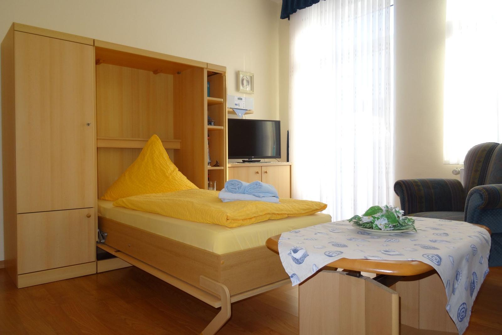 Schlafzimmer mit Bett, Fernseher und Tisch. Fenster mit Vorhängen.