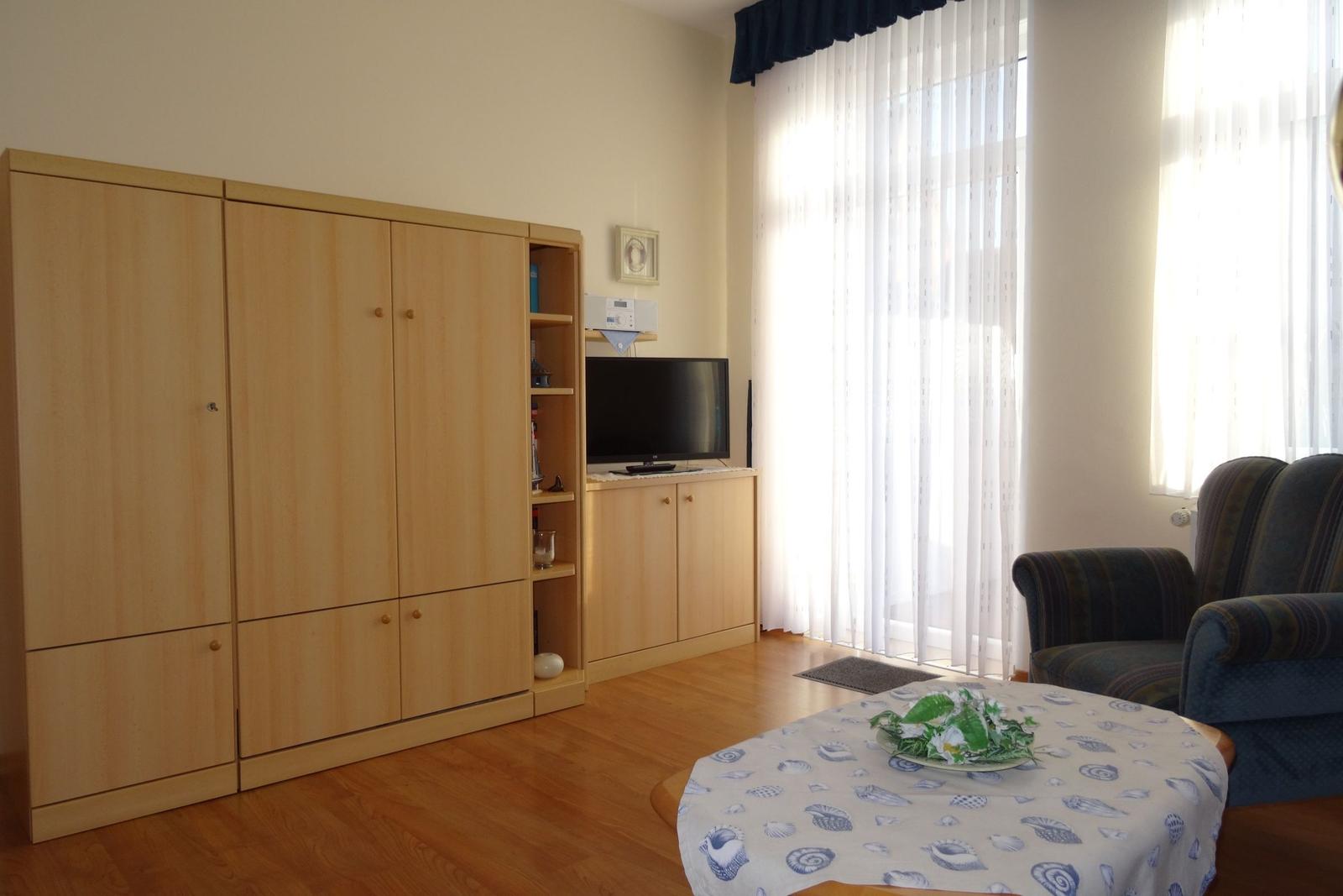 Wohnzimmer mit Holzmöbeln, Fernseher und Sitzgelegenheit. Tisch mit Tischdecke und Blumen.