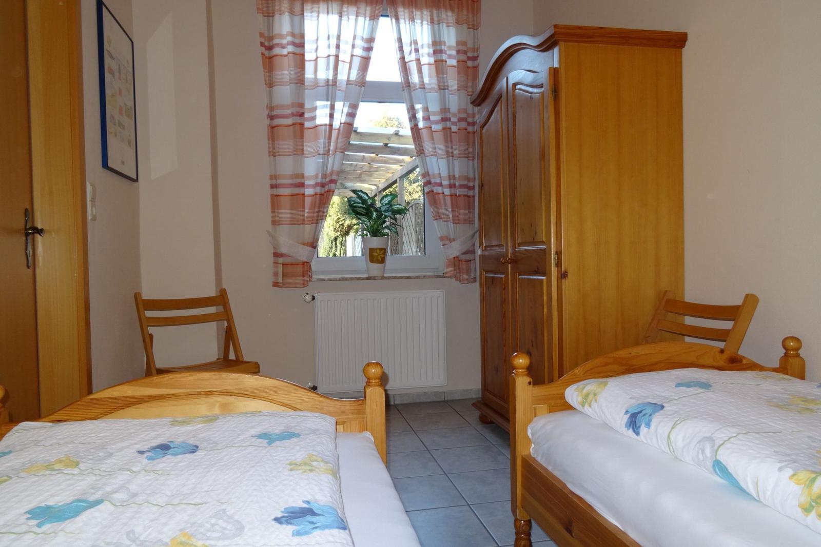 Zwei Betten, Schrank und Fenster mit Vorhängen. Draußen ist eine Terrasse mit Pflanzen sichtbar.