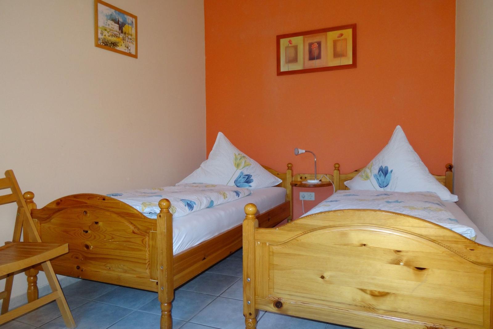 Zwei Einzelbetten mit Holzgestell, Wand mit Orange und Bilder, Nachttisch mit Lampe.