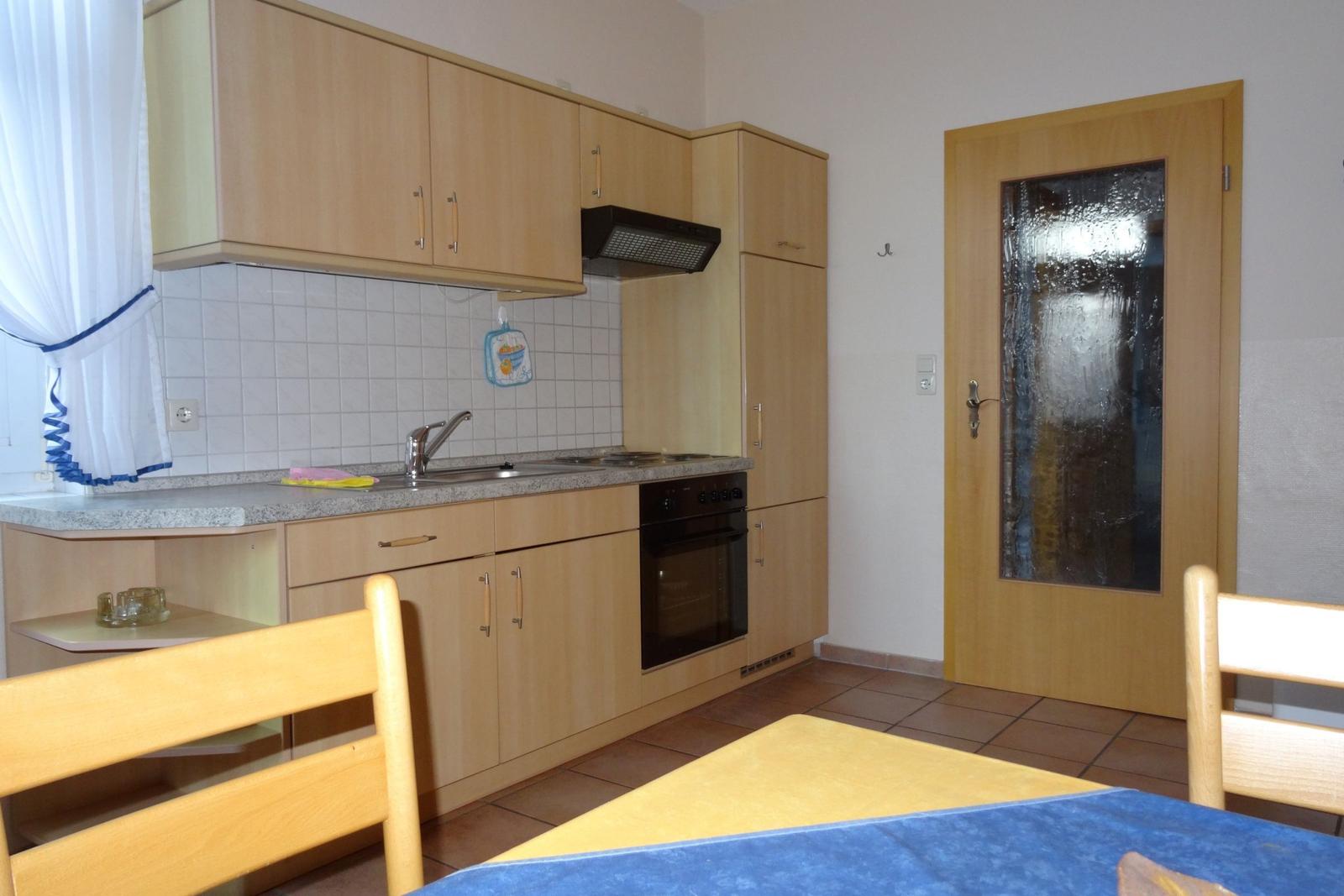 Küche mit Holzschränken, Spüle und Ofen. Tür mit Glasfenster. Esstisch mit Stühlen.