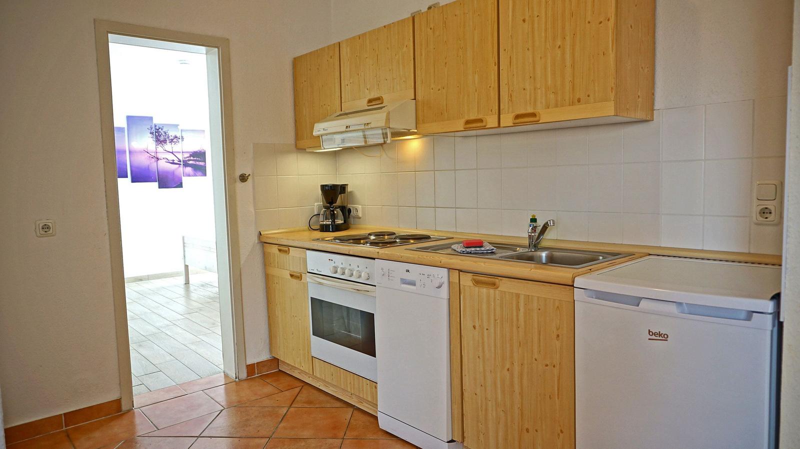 Küche mit Holzschränken, Spüle, Herd und Kühlschrank. Tür führt in einen Flur mit Wandbild.