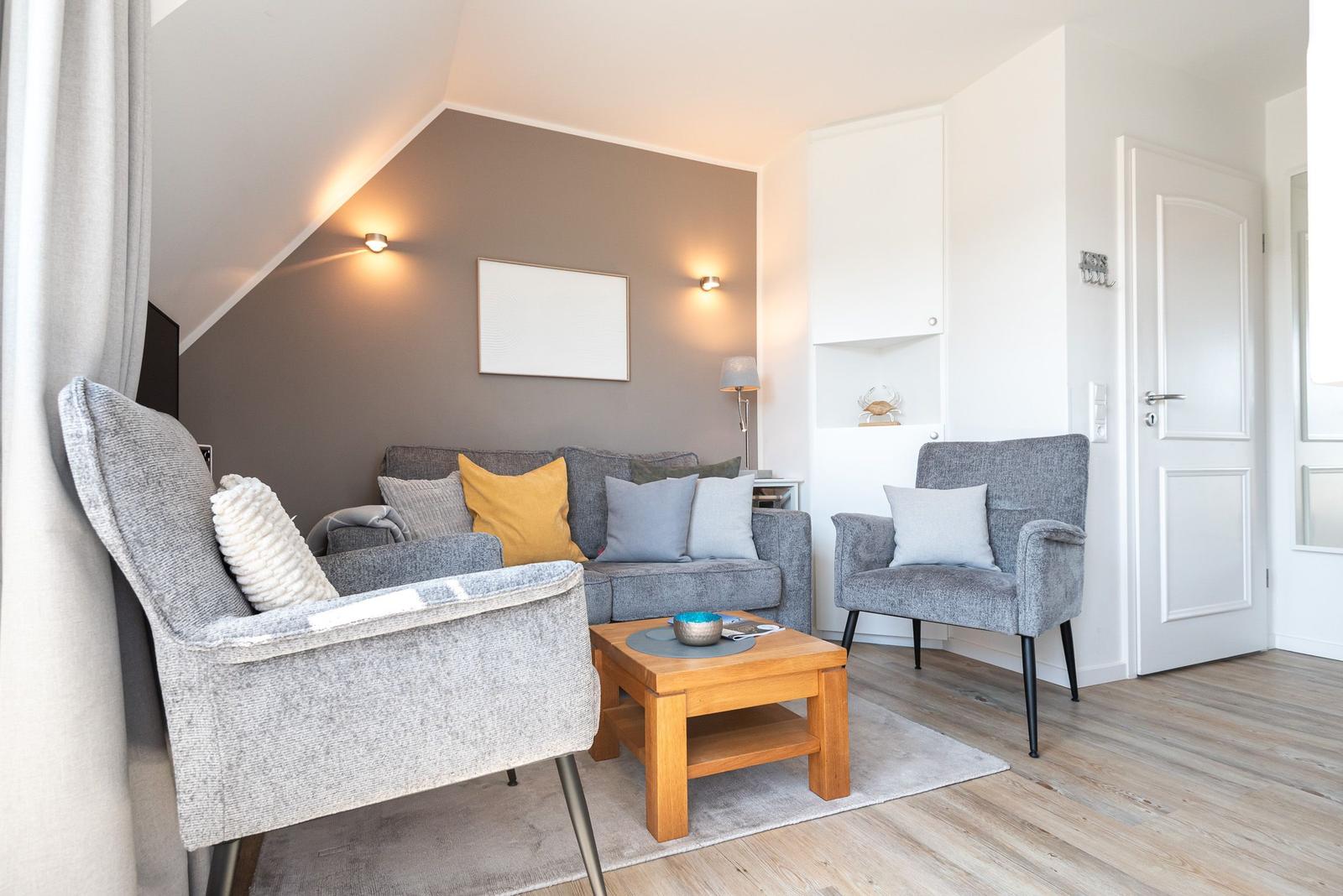 Wohnzimmer mit grauer Couch, zwei Sesseln und Holztisch. Wandfarbe grau, Holzboden und weiße Türen.