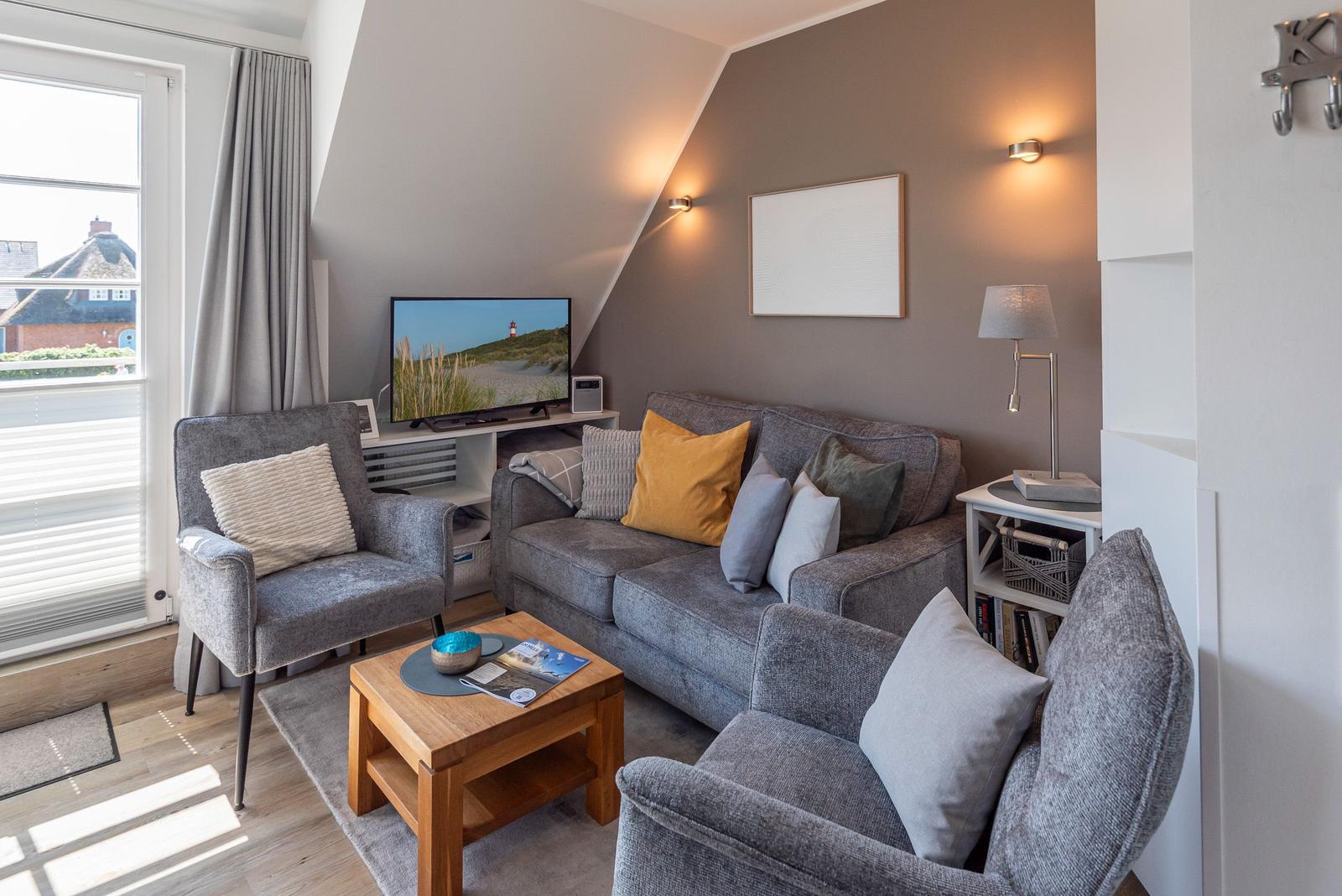 Wohnzimmer mit Sofa, Sessel und Fernseher. Fenster mit Blick auf Dachhäuser.