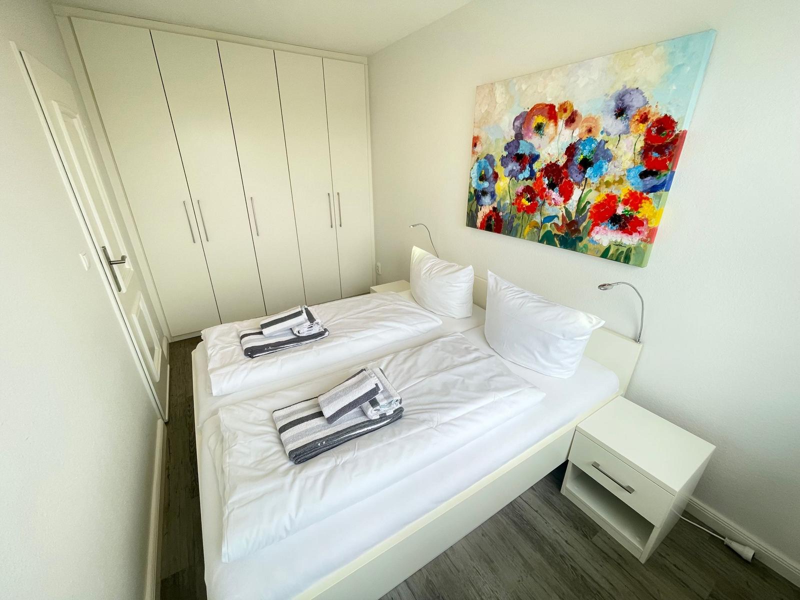 Schlafzimmer mit Doppelbett, weißen Möbeln und Blumenbild an der Wand.