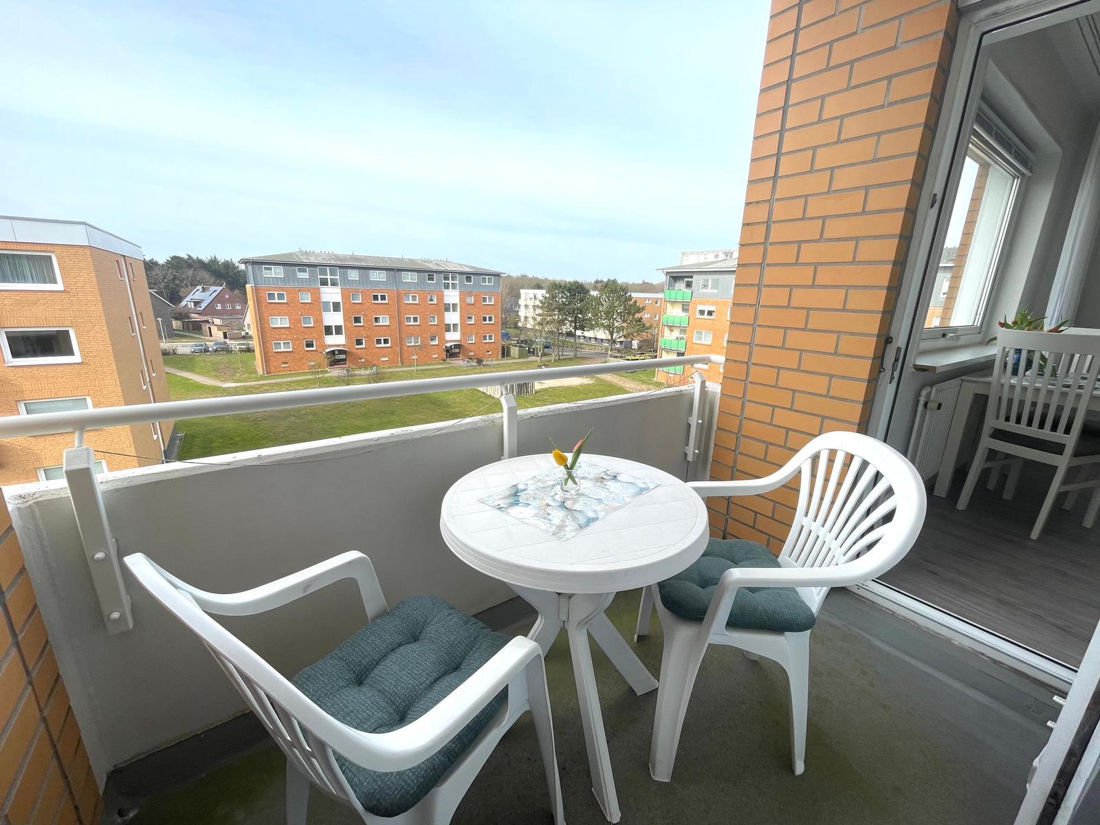 Balkon mit weißem Tisch und zwei Stühlen. Blick auf Wohnhäuser und Grünflächen.