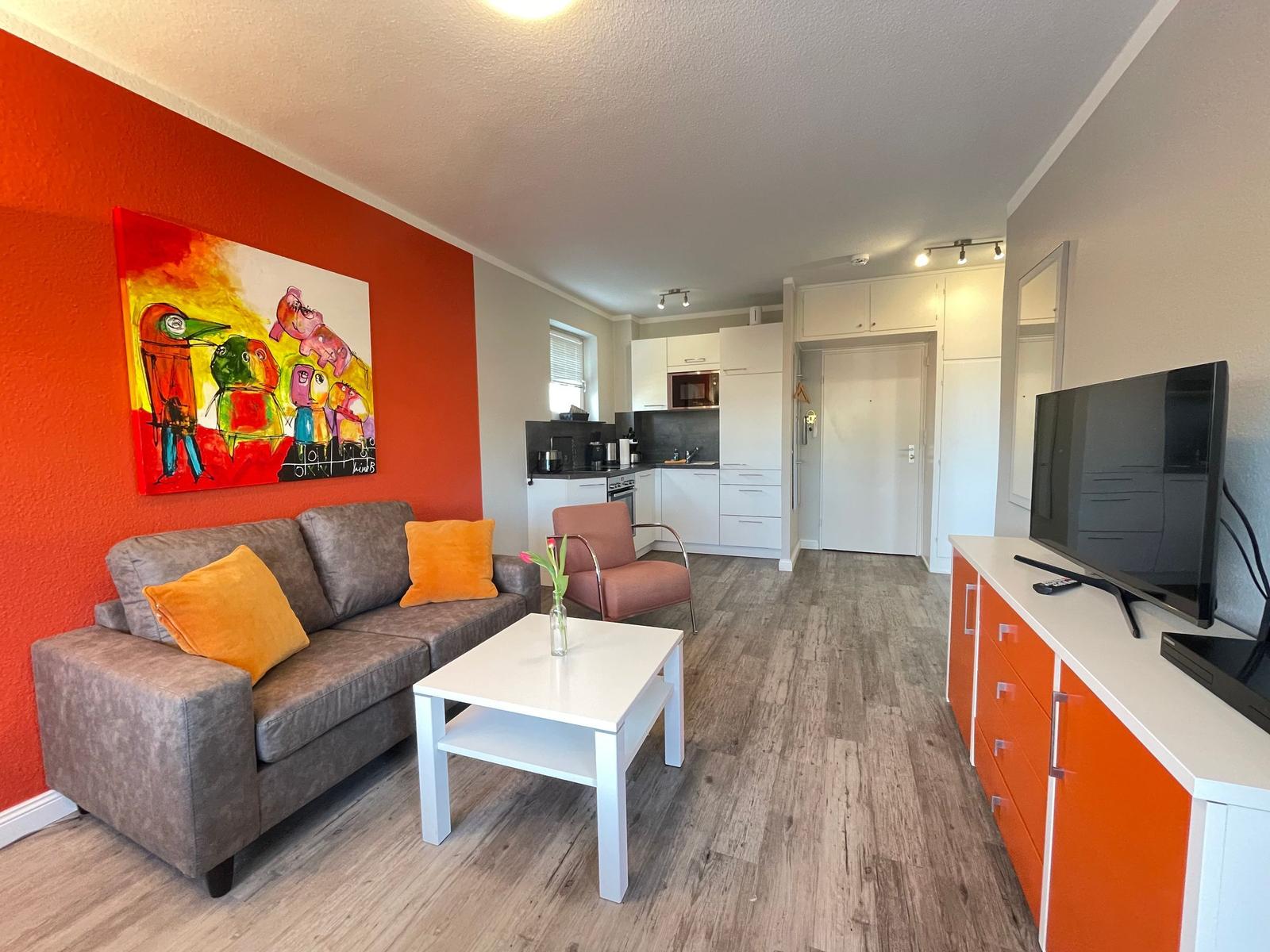 Wohnbereich mit Sofa, Couchtisch und Fernseher. Hintergrund: Küche und orange Wand mit Kunstwerk.