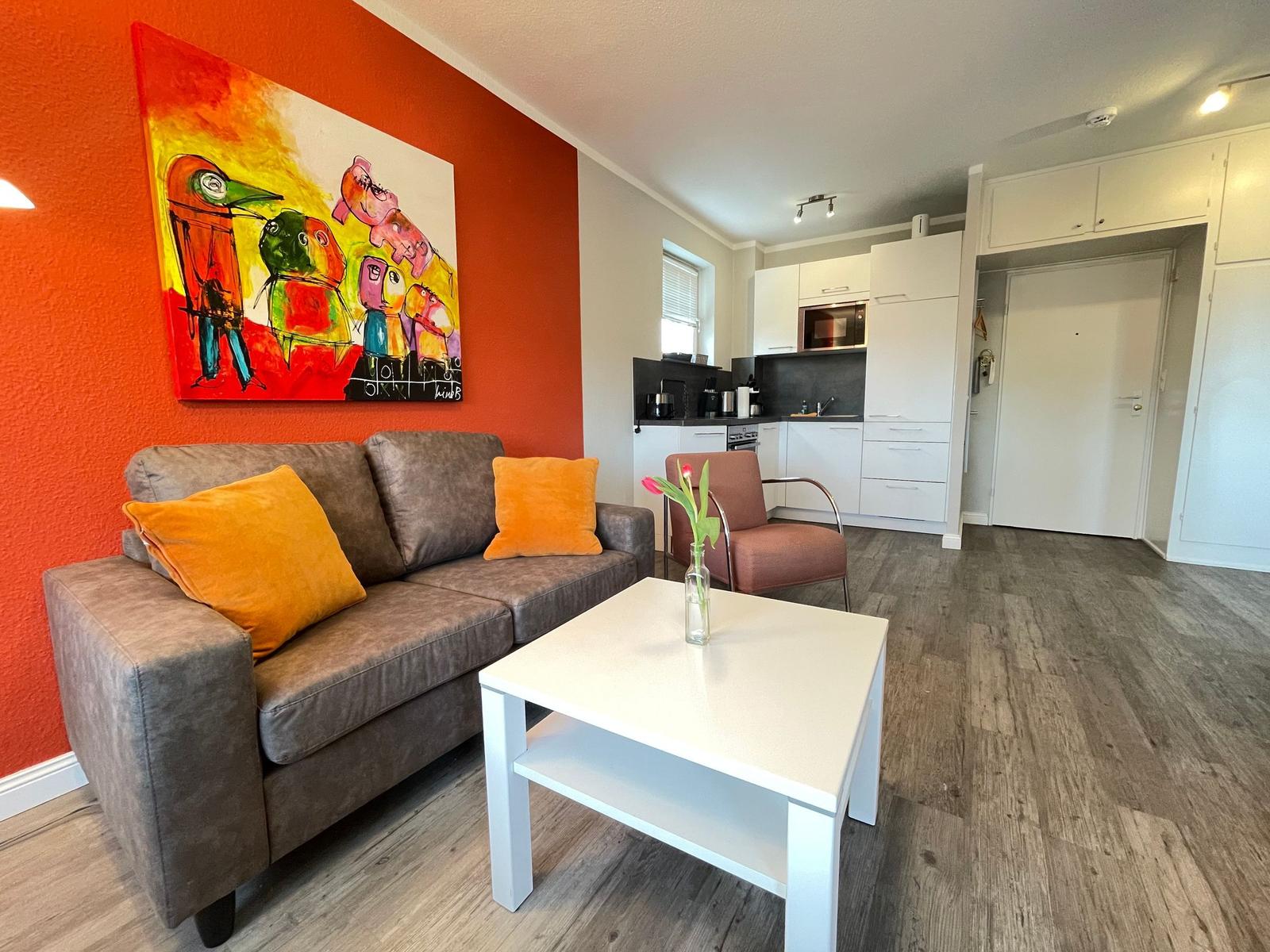 Wohnzimmer mit Sofa, Kaffeetisch und Wandbild. Küche und Flur sind sichtbar.