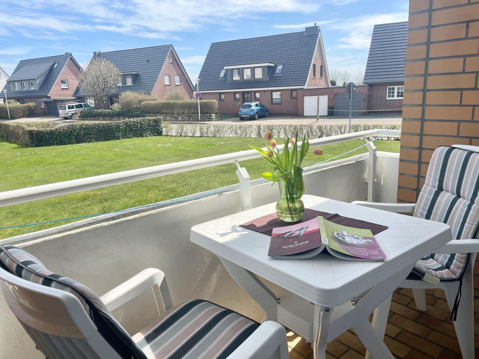 Balkon mit Tisch, zwei Stühlen und Blumen. Blick auf Gras und Häuser.