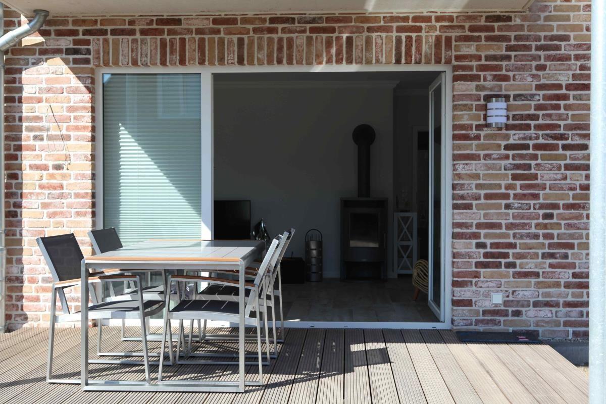Terrasse mit Tisch und Stühlen. Durch Glastür ist Wohnbereich mit Kamin sichtbar.