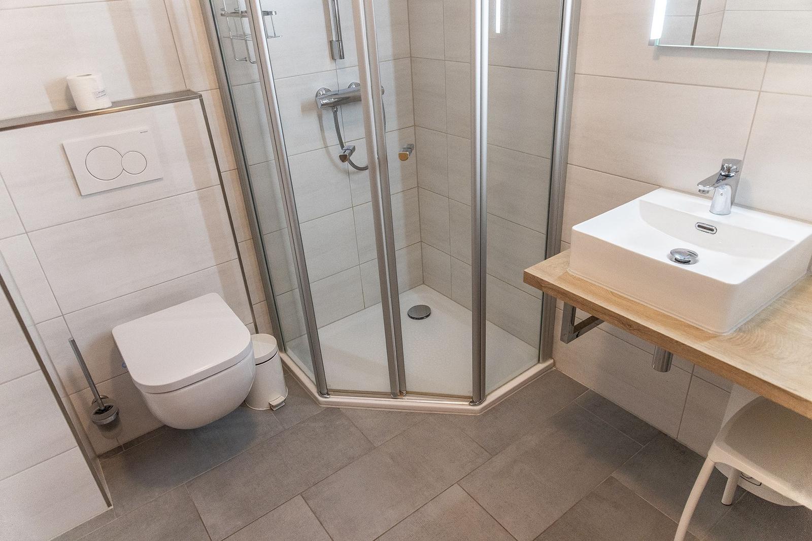 Badezimmer mit Dusche, Toilette und Waschbecken auf Holzunterlage.