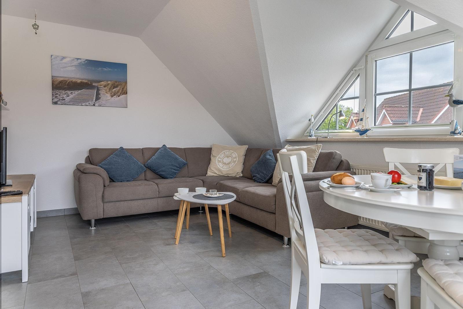 Wohn- und Essbereich mit Sofa, Tisch und Fenster. Wandbild und Küchenecke sichtbar.