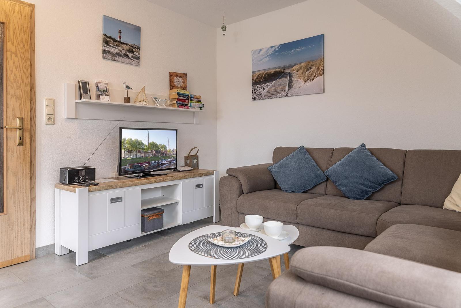 Wohnzimmer mit Sofa, Fernseher und Tisch. Wandbilder zeigen Strand und Leuchtturm.