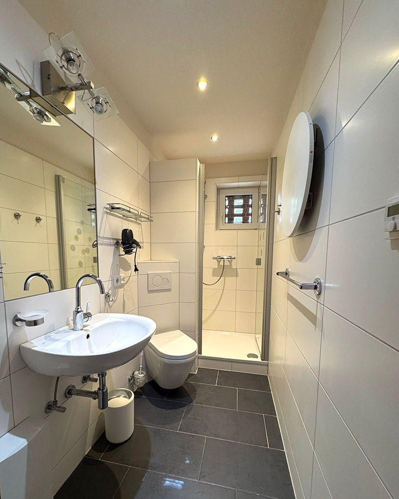 Badezimmer mit Dusche, Waschbecken und WC. Weiße Fliesen und dunkle Bodenfliesen.