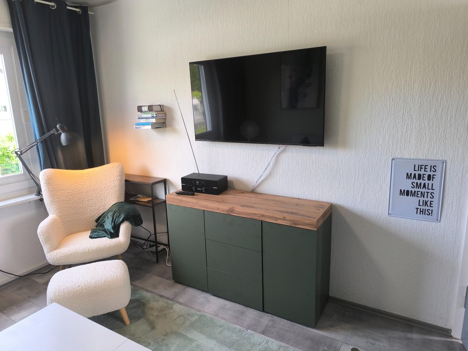 Wohnzimmer mit Fernseher, grünem Schrank und weißem Sessel.