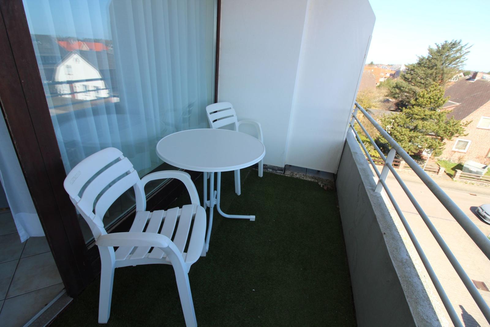 Balkon mit weißem Tisch und zwei Stühlen, grüner Teppich, Blick auf Wohngebiet.