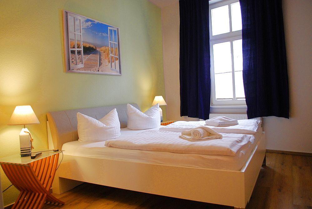 Schlafzimmer mit Bett, Nachttisch, Lampe und Fenster mit Vorhängen.