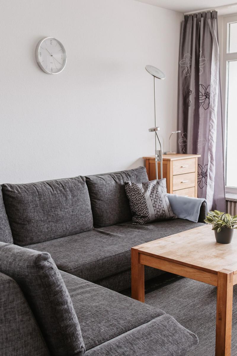 Ein Wohnzimmer mit grauem Sofa, Holztisch und Vorhängen an einem Fenster.
