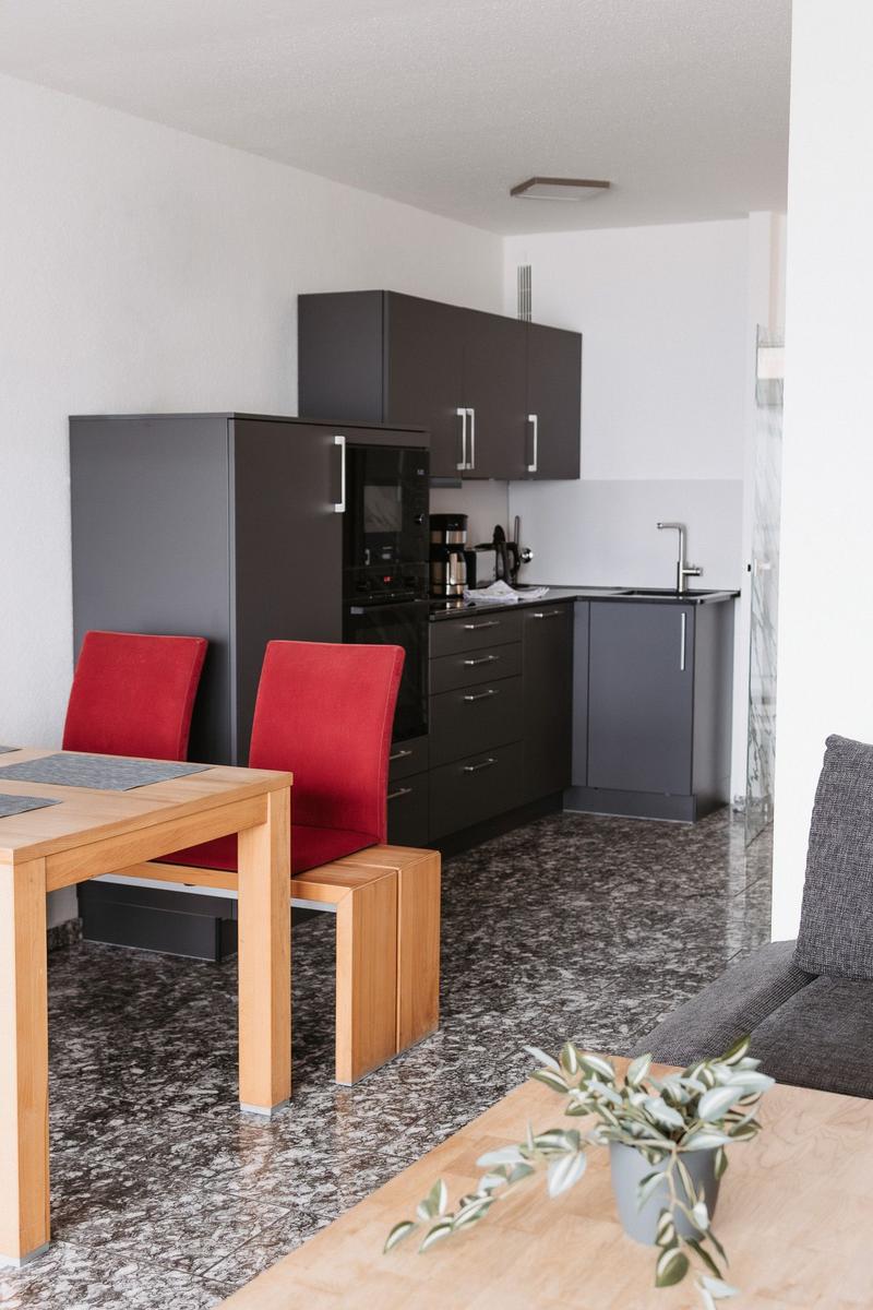 Küche mit dunklen Schränken, Essbereich mit roten Stühlen und Wohnbereich mit grauem Sofa.