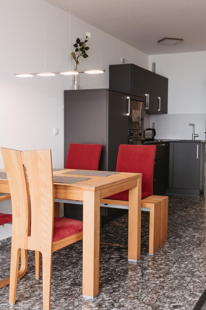 Essbereich mit Holztisch, roten Stühlen und dunkler Küchenzeile im Hintergrund.